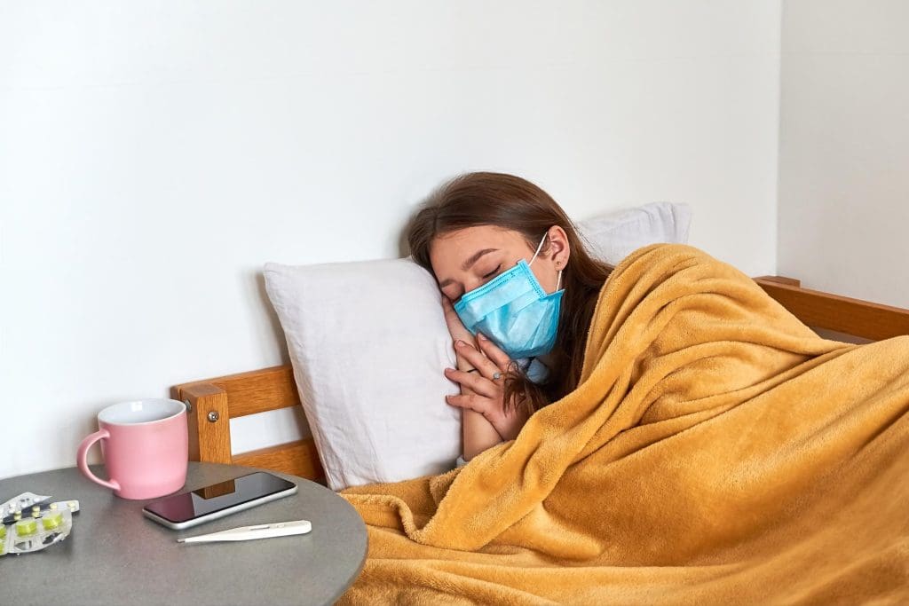 Sick Girl with Flu with Protective Mask on Face Laying Under Blanket in Bed at Home. Pills Thermometer and Tablets on Table.